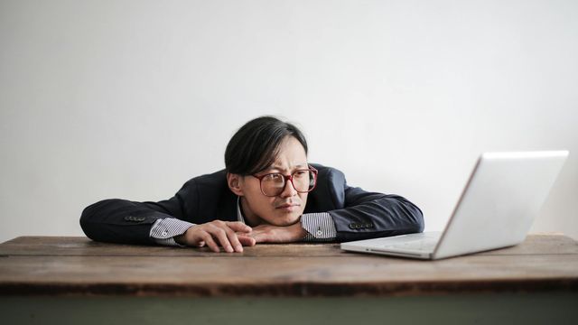 A man sits slumped at a desk looking at a laptop. 