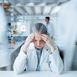 A stressed scientist sits at a lab bench with her head in her hands as other scientists pass, blurred in the background. 