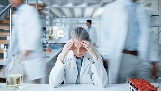 A stressed scientist sits at a lab bench with her head in her hands as other scientists pass, blurred in the background. 