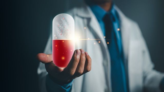 Scientist in a lab coat holding a large red and white drug capsule. Inside the drug is a molecular structure and yellow futuristic-looking dots. 