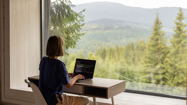 A woman sits at a desk working on a laptop in front of a large window. 